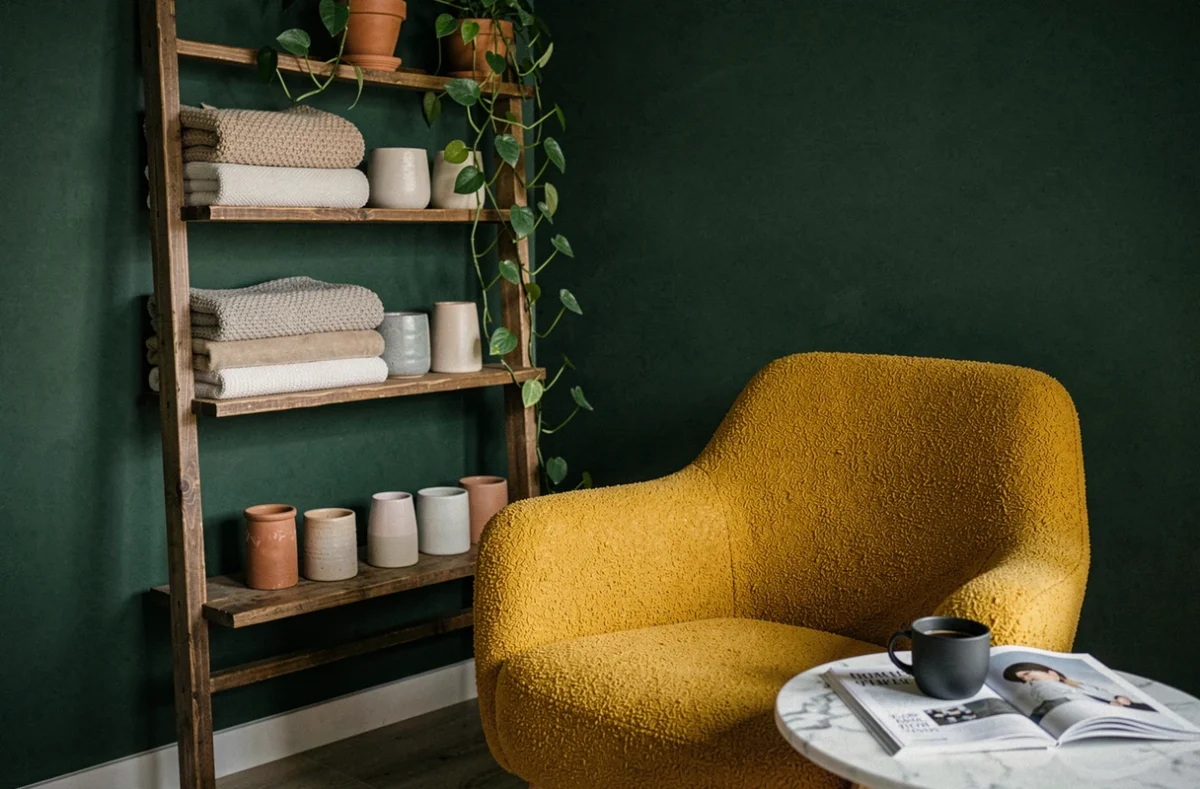 Coin salon décoré avec un mur vert forêt, une étagère en bois vintage et un fauteuil en bouclé moutarde avec des accessoires en céramique